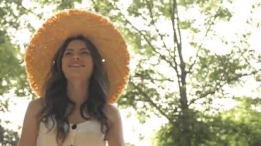 Hasır şapkalı, mutlu, çekici, esmer bir kadının parkta yürürken gülümsemesi ve dönmesi.