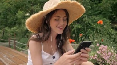 Hasır şapkalı, hoş, genç, esmer bir kadın. Kulaklıklı müzik dinliyor ve parkta bankta otururken akıllı telefondan sohbet ediyor.