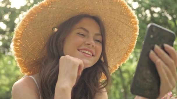 Hasır Şapkalı Mutlu Genç Esmer Bir Kadın Parkta Otururken Telefon — Stok video