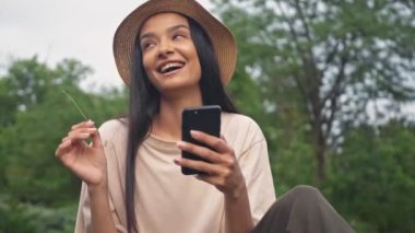 Parkta akıllı telefon kullanırken şapkalı neşeli, esmer Asyalı kadın elinde samanla gülüyor ve oynuyor.