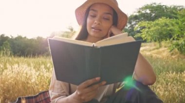 Parkta ekose üzerinde otururken şapkalı, gülümseyen ve kitap okuyan tatlı, sakin, genç esmer Asyalı kadın.