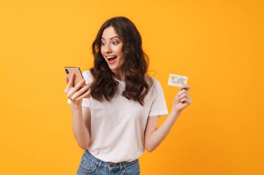 Cep telefonu banka kartı tutarak sarı duvar arka plan üzerinde izole poz şok genç kadın.