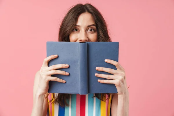 Mooie gelukkige emotionele jonge schattige vrouw poseren geïsoleerd over roze muur achtergrond dekking gezicht met boek. — Stockfoto