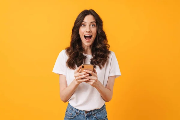 Pleased shocked young woman posing isolated over yellow wall background using mobile phone. — Stock Photo, Image