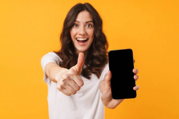 Glad positiv leende ung kvinna poserar isolerade över gul vägg bakgrund med hjälp av mobiltelefon som visar displayen och tummen upp. — Stockfoto