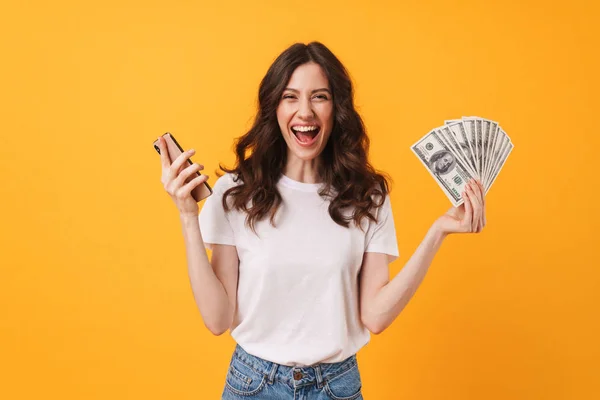 Plaisir heureux souriant jeune femme posant isolé sur fond de mur jaune en utilisant un téléphone portable tenant de l'argent . — Photo