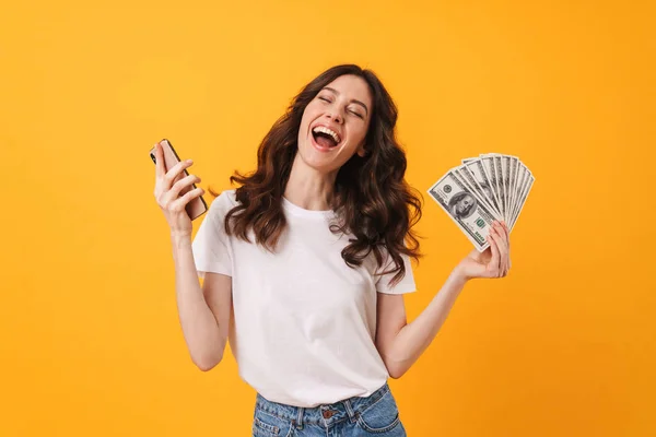 Blij gelukkig glimlachend jonge vrouw poseren geïsoleerd over gele muur achtergrond met behulp van mobiele telefoon houden van geld. — Stockfoto