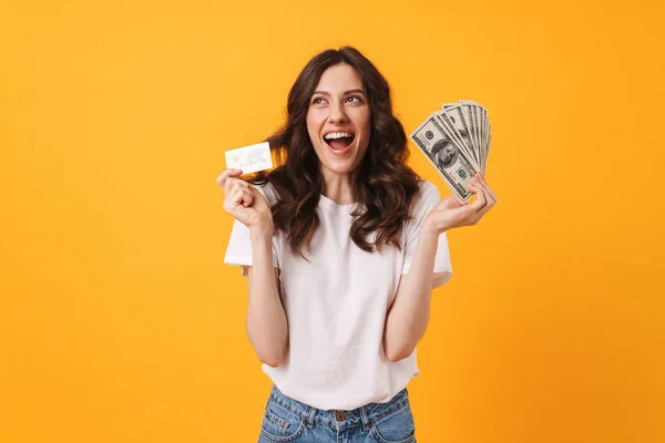 Agradable feliz sonriente joven posando aislado sobre fondo de pared amarilla celebración de dinero y tarjeta de débito . — Foto de Stock