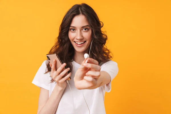 Mujer joven posando aislada sobre fondo amarillo de la pared escuchando música usando el teléfono móvil le dan un auricular . — Foto de Stock