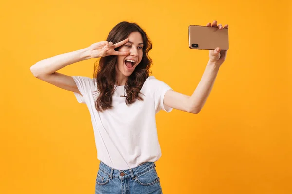 Positive optimistic smiling young woman posing isolated over yellow wall background take a selfie by mobile phone. — Stock Photo, Image