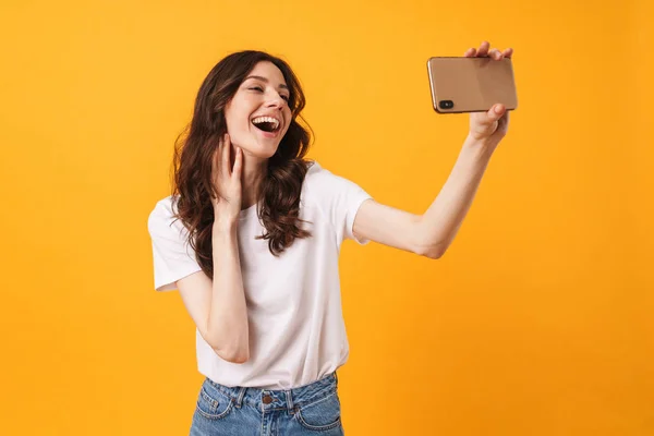 Positiv optimistisk leende ung kvinna poserar isolerade över gul vägg bakgrund ta en selfie med mobiltelefon. — Stockfoto