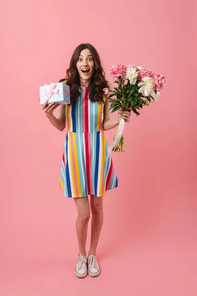 Досить здивована позитивна мила жінка позує ізольовано на фоні рожевих стін, тримаючи подарункову коробку та квіти . — стокове фото