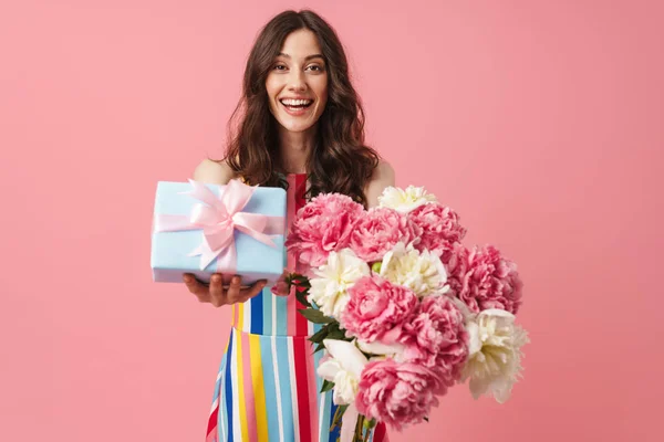 Pěkná, překvapena, milá žena, která pózuje na pozadí růžové zdi a drží dárek s dárkem a květinami. — Stock fotografie