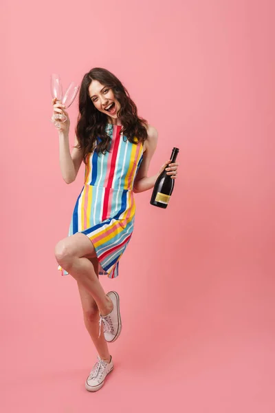 Bastante contenta mujer linda positiva posando aislada sobre fondo de pared rosa sosteniendo vasos y botella con champán . — Foto de Stock