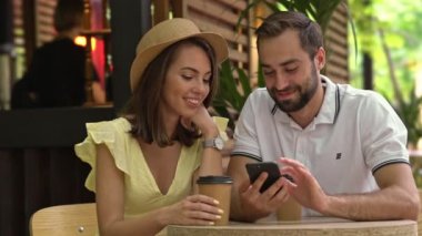 Güzel gülümseyen genç bir çift kafede otururken konuşuyor ve akıllı telefon kullanıyor.