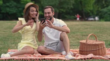 Heyecanlı genç çift akıllı telefonlarıyla müzik dinliyor ve parkta ekose üzerinde otururken şarkı söylüyor.