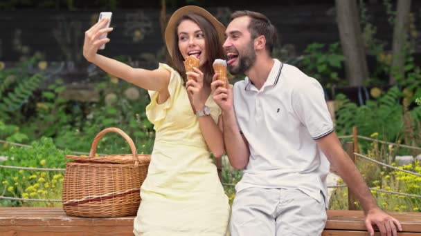 快乐的年轻夫妇坐在公园的长椅上 一边吃着冰淇淋 一边用智能手机拍着自己的照片 — 图库视频影像