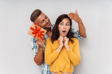 Gülümseyen neşeli adam gri duvar arka plan üzerinde izole onun şok kadın bir hediye vermek.