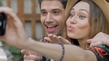 Gülümseyen genç çiftin yakın görüntüsü. Akıllı telefondan selfie çekerken elleriyle göz kırpan ve el kol hareketi yapan çiftin.