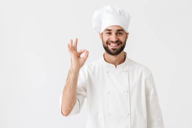 Gülümseyen genç aşçı, beyaz duvar arkasında yalnız poz veriyor. Üniformayla iyi bir hareket gösteriyor..