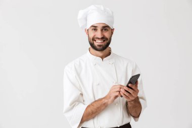 Neşeli gülümseyen genç aşçı üniformalı beyaz duvar arkasında cep telefonu kullanarak poz veriyor..