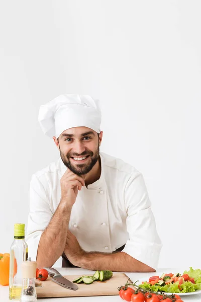 Nöjd positiv glad ung kock isolerad över vit vägg bakgrund i enhetlig matlagning med färska grönsaker. — Stockfoto