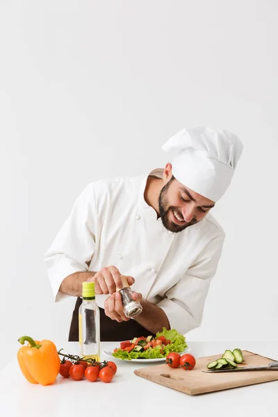 Ευχαριστημένος χαρούμενος νεαρός σεφ απομονωμένος σε λευκό φόντο τοίχου σε ομοιόμορφο μαγείρεμα με φρέσκα λαχανικά. — Φωτογραφία Αρχείου