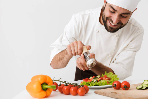 Photo of cheery pleased young chef isolated over white wall background in uniform cooking with fresh vegetables.
