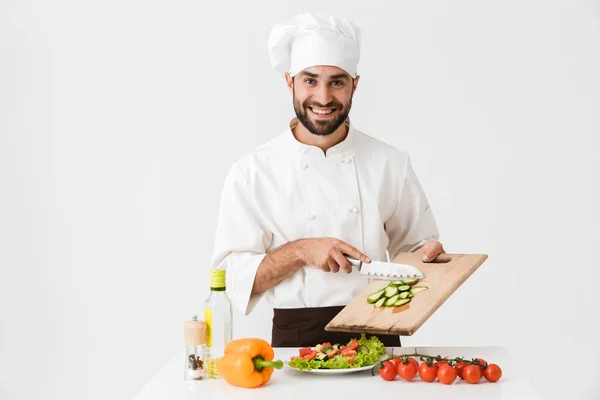 Imagem de homem cozinheiro alegre em uniforme sorrindo e cortando vegeta — Fotografia de Stock