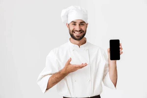 Positif souriant jeune chef posant isolé sur fond de mur blanc en uniforme tenant téléphone portable montrant affichage vide . — Photo