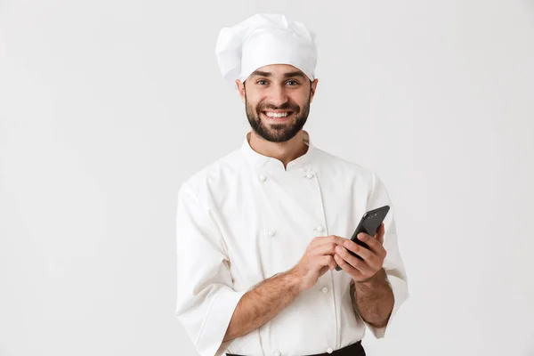 Wesoły uśmiech młody szef kuchni pozowanie na białym tle ściany w mundurze za pomocą telefonu komórkowego. — Zdjęcie stockowe