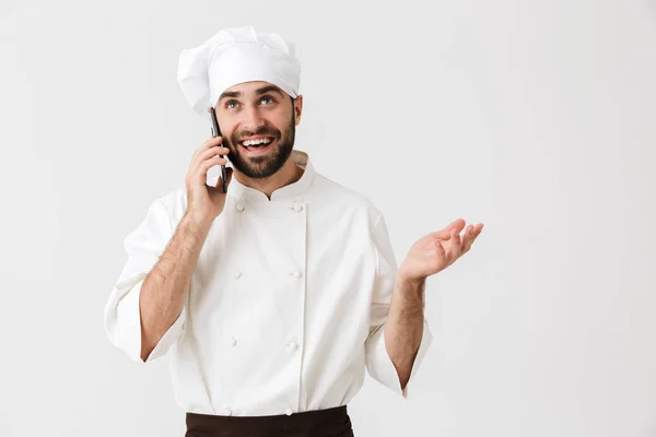 Эмоциональный счастливый молодой шеф-повар позирует изолированно на фоне белой стены в униформе, разговаривая по мобильному телефону . — стоковое фото