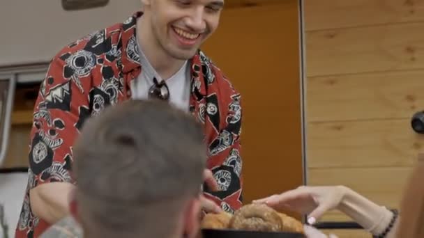 Beau jeune homme brunet joyeux sortir de la remorque avec des croissants chauds et rejoindre ses amis à la table en bois tout en s'amusant ensemble au pique-nique 