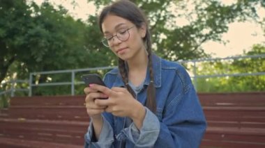 Kot ceketli tatlı, mutlu, esmer kız kaykayının yanında otururken gülümsüyor ve akıllı telefon kullanıyor.