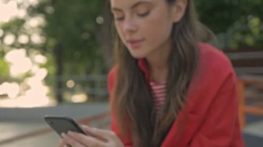Parkta otururken kırmızı ceketli, gülümseyen ve akıllı telefondan sohbet eden mutlu genç kızın yakın görüntüsü.