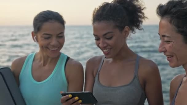 海の近くにいるスマートフォンで何かを見ている間にスポーツウェアで話して笑っている若いスポーティ多民族の女性の友人のグループ — ストック動画