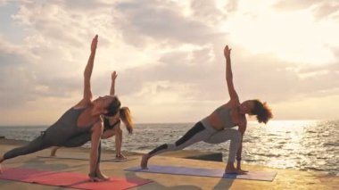 Spor kıyafetleri giymiş, deniz kenarındaki minderler üzerinde yoga yapan bir grup konsantre genç, sportif, etnik çeşitlilikte kadın arkadaş.