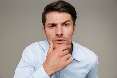 Resmi kıyafetler giyen, kameraya bakıp çenesine dokunan düşüncelere dalmış ciddi bir adamın görüntüsü.