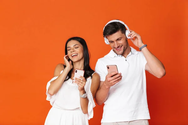 Uśmiechnięta młoda niesamowita kochająca się para pozująca na czerwonym tle ściany za pomocą telefonów komórkowych słuchających muzyki ze słuchawkami. — Zdjęcie stockowe