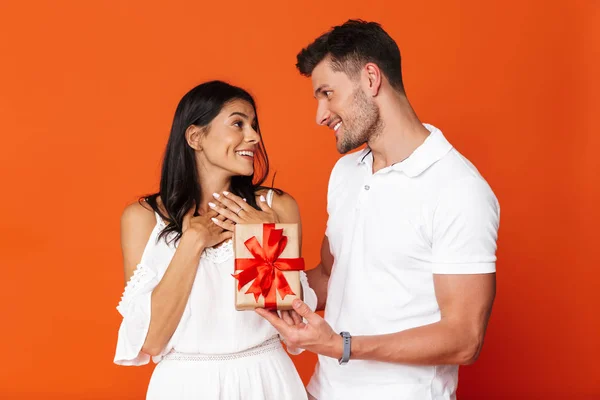 Alegre feliz complacido joven dar un regalo sorpresa a su esposa novia aislado sobre fondo de la pared roja . — Foto de Stock
