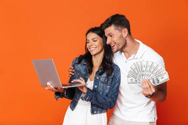 Alegre casal amoroso otimista emocional isolado sobre fundo parede vermelha segurando dinheiro usando computador portátil . — Fotografia de Stock