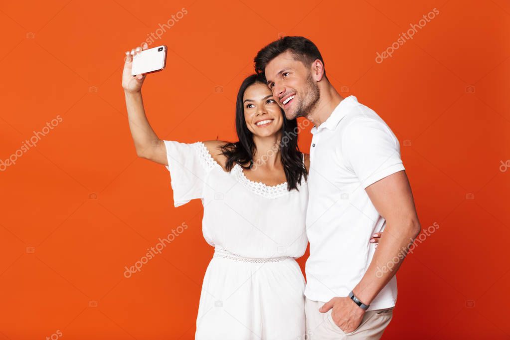 Happy positive pleased young amazing loving couple posing isolated over red wall background take selfie by mobile phones.