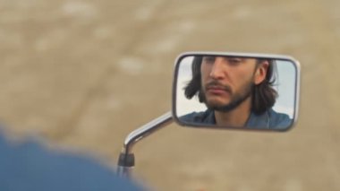 Yakışıklı, esmer, sakallı bir adamın sahilde motosiklette otururken aynaya bakıp saçını düzeltmesi.