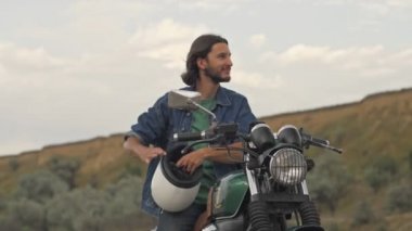 Yakışıklı, esmer, sakallı adam kaskını takıyor ve plajda motosikletin üstünde otururken motoru çalıştırıyor.