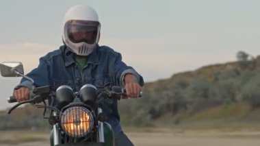 Kasklı, ciddi bir genç adam motosikletinin başında oturuyor ve plajda arabayla uzaklaşıyor.