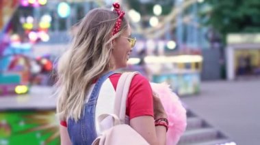 Eğlence parkında yürüyen ve pamuk helva yemekten hoşlanan neşeli sarışın bir kadının yan görüntüsü.