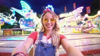 Çekici, mutlu, genç, sarışın bir kadın lunaparkta fotoğraf çekerken yüzünü buruşturuyor ve eğleniyor.