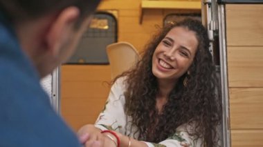 Çekici, genç esmer, kıvırcık saçlı, karavanın yanında oturmuş erkek arkadaşıyla gülümseyen ve konuşan kadın.