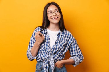 İyimser, mutlu, gözlüklü genç kız sarı duvar arkasında izole bir şekilde poz veriyor..
