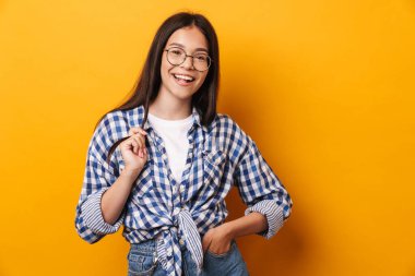 İyimser, mutlu, gözlüklü genç kız sarı duvar arkasında izole bir şekilde poz veriyor..